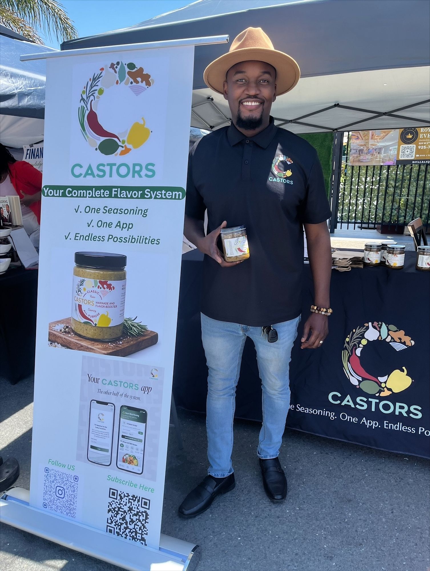 A CASTORS team member smiling at an outdoor market booth with a branded banner and product jars